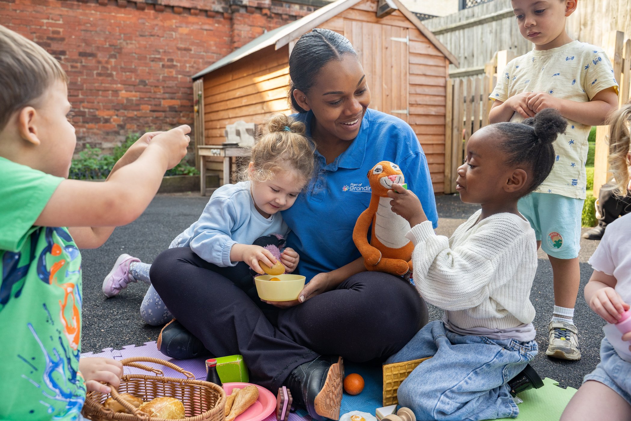 Diverse children gather around a smiling caregiver outdoors, sharing snacks and a plush toy in a warm, joyful scene of inclusive play.