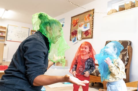 Adult and two children play indoors wearing translucent green and blue veils, smiling and laughing in a warm, playful family scene.