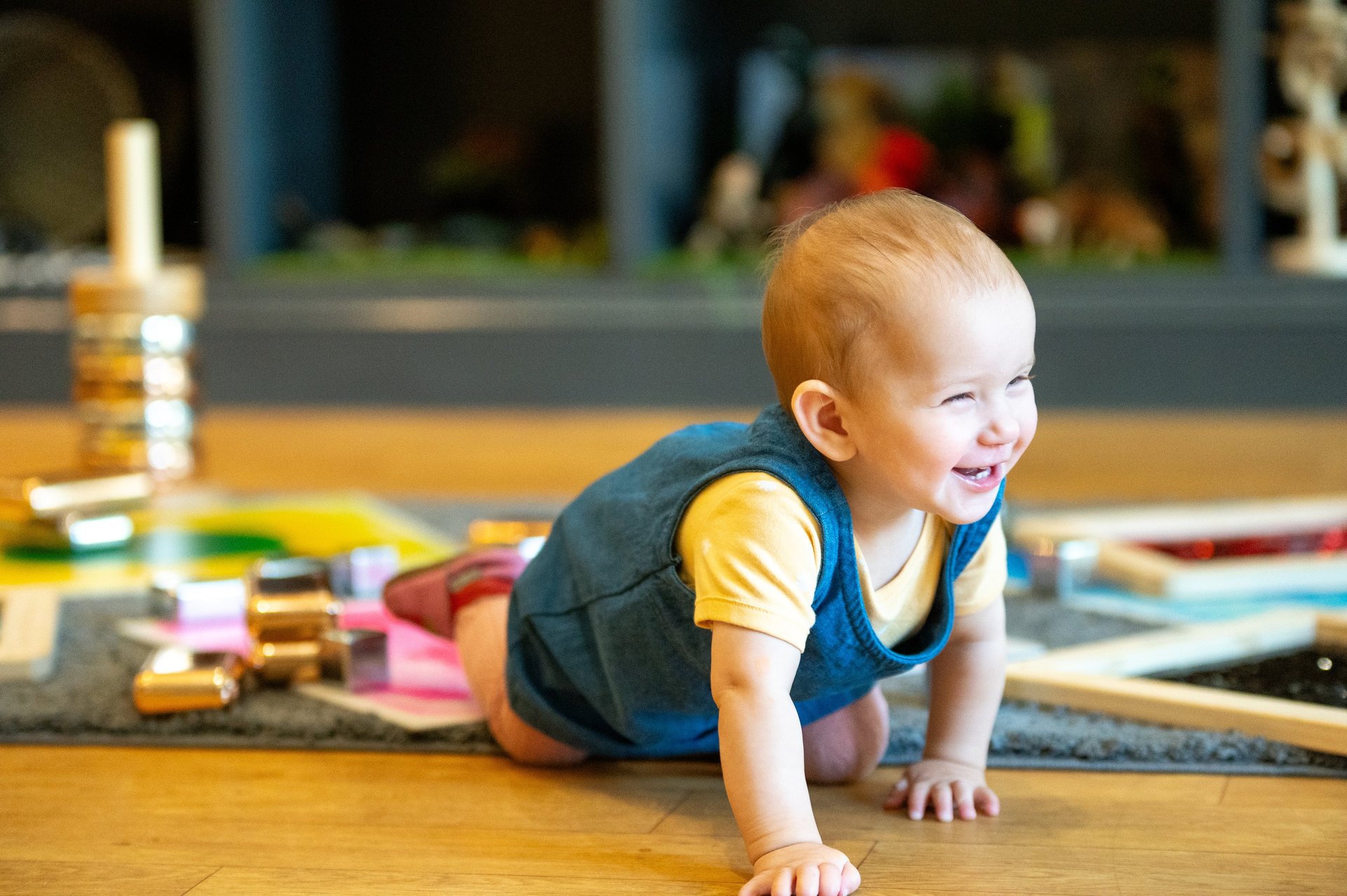 Smiling baby in a yellow shirt and dark vest crawls across a wooden floor in a bright playroom, with toys and books blurred behind.