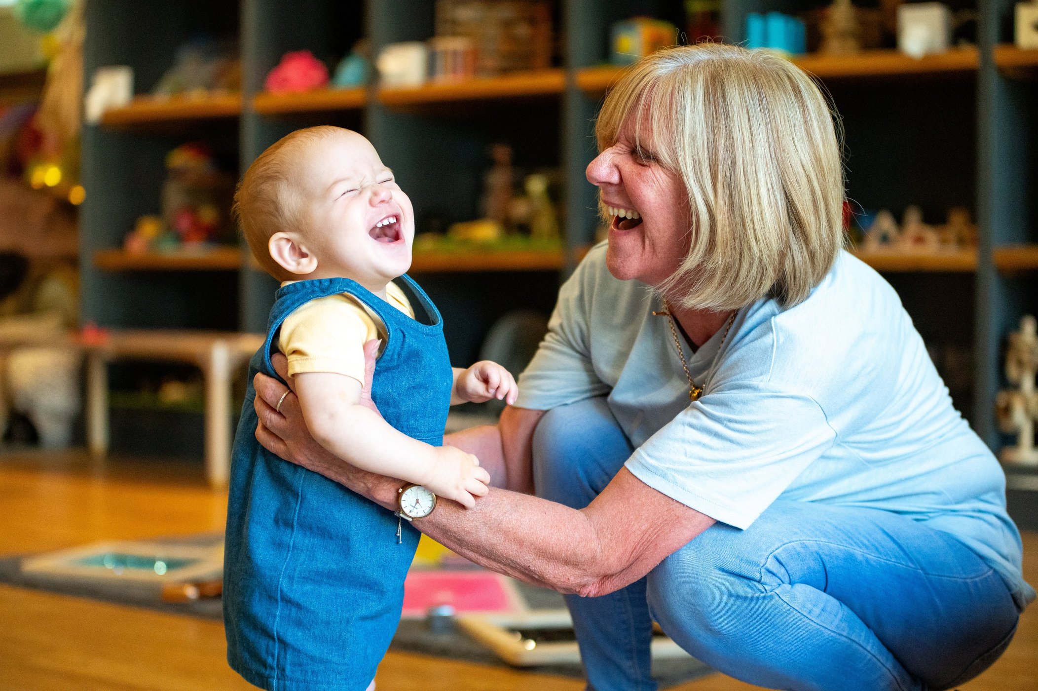 Grandmother laughing with a joyful baby in denim overalls as they play together in a colourful room filled with shelves and toys.