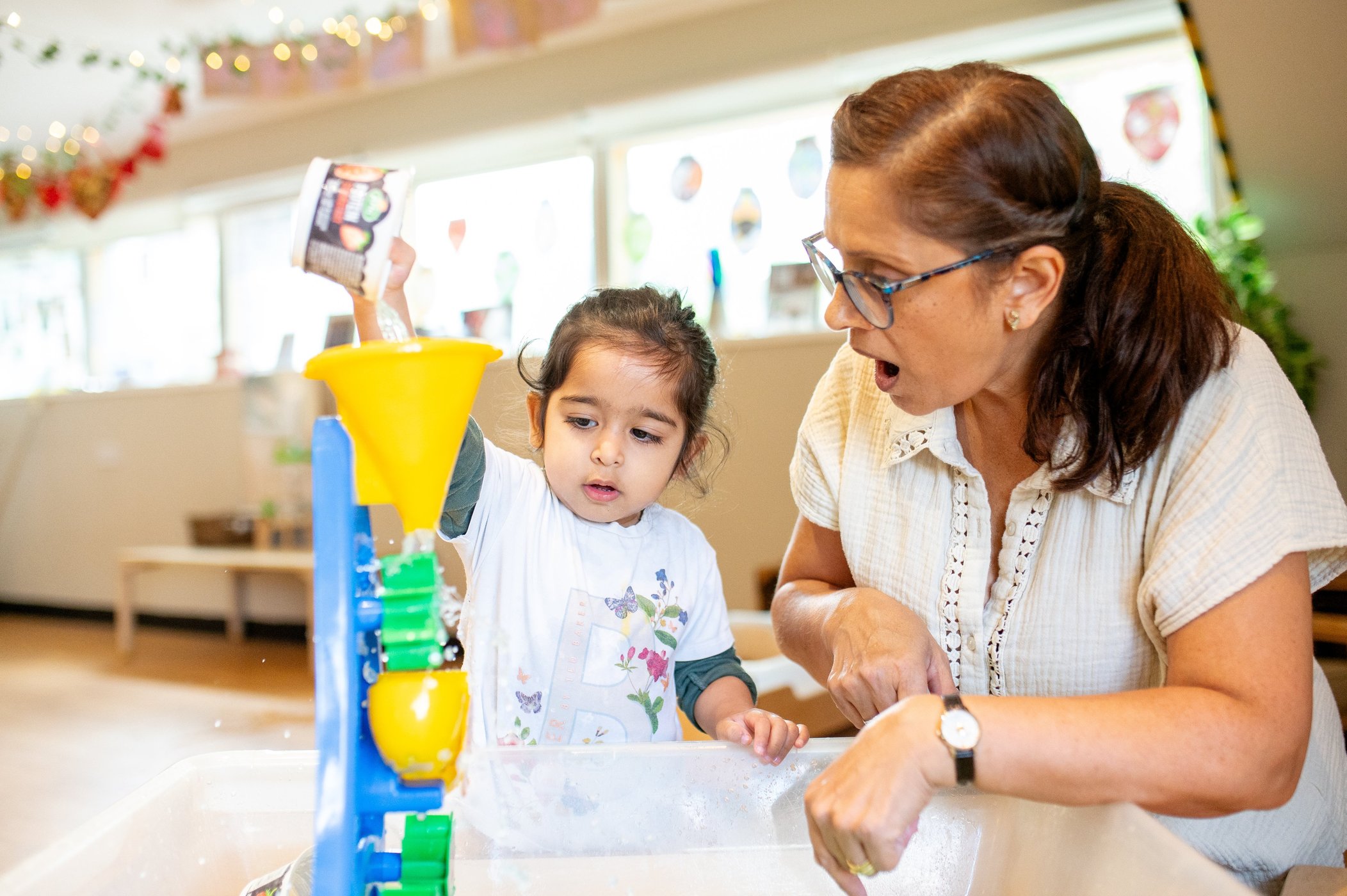 Woman and young girl play with a colourful water table indoors; the girl pours into a yellow funnel while the woman guides her, engaged.