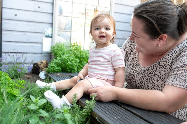 Smiling toddler in a pink striped outfit sits on a garden bench as a woman embraces her, surrounded by flowers and lush greenery.
