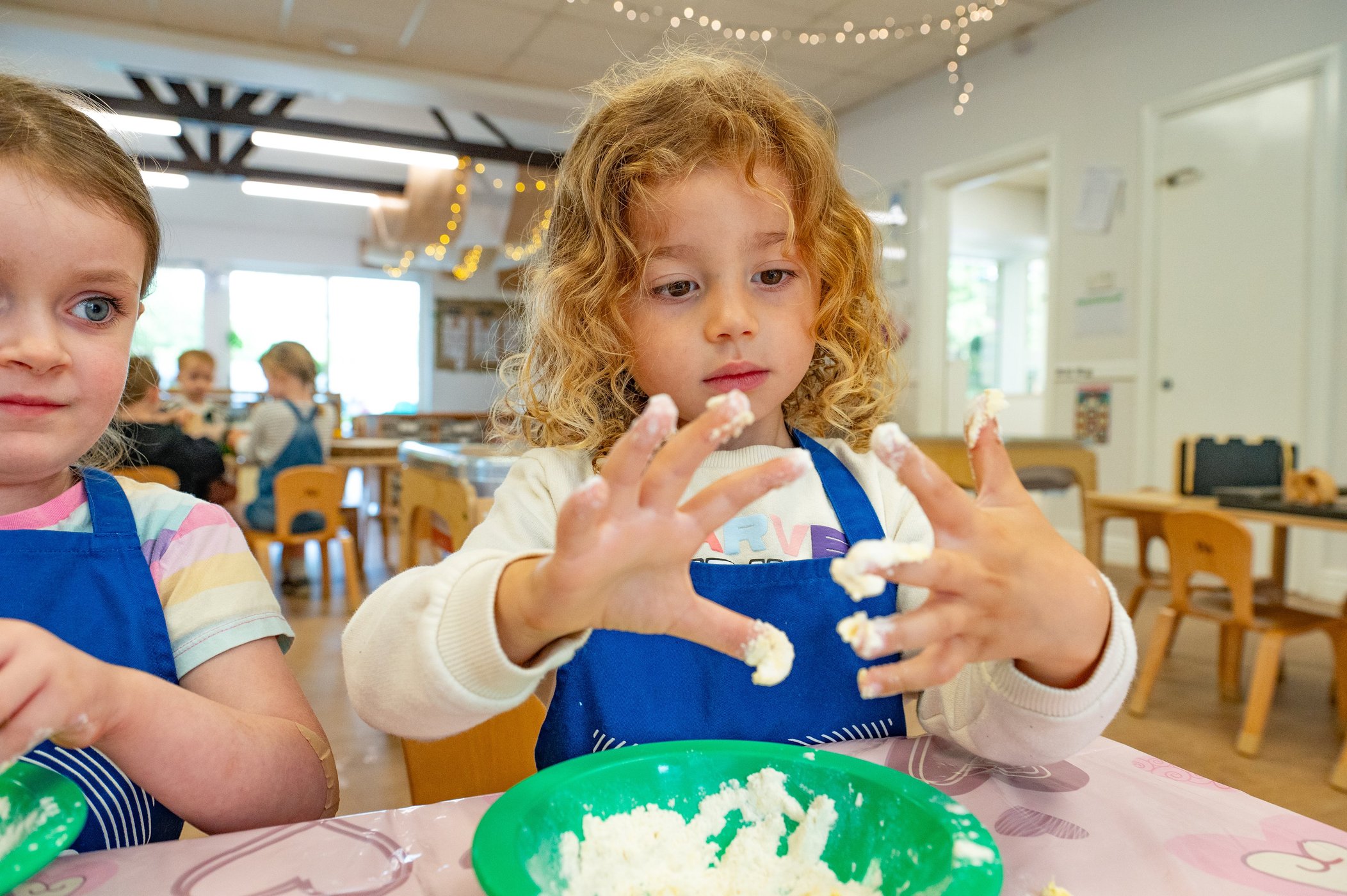 Young girl in a blue apron molds white dough at a classroom table, focused, while another child watches in a bright, cheerful room.