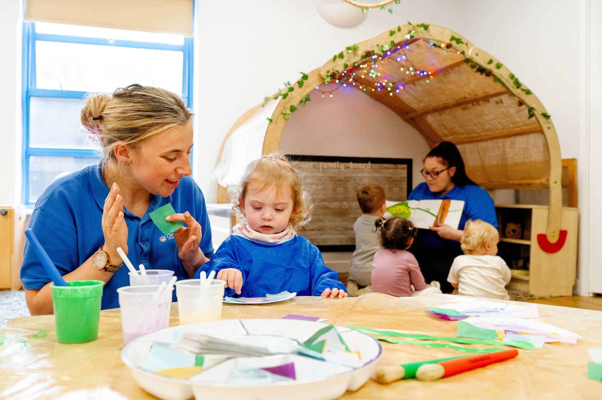 In a bright classroom, a teacher helps a toddler paint as children play in a decorated tent; art supplies fill the table in a warm, joyful scene.