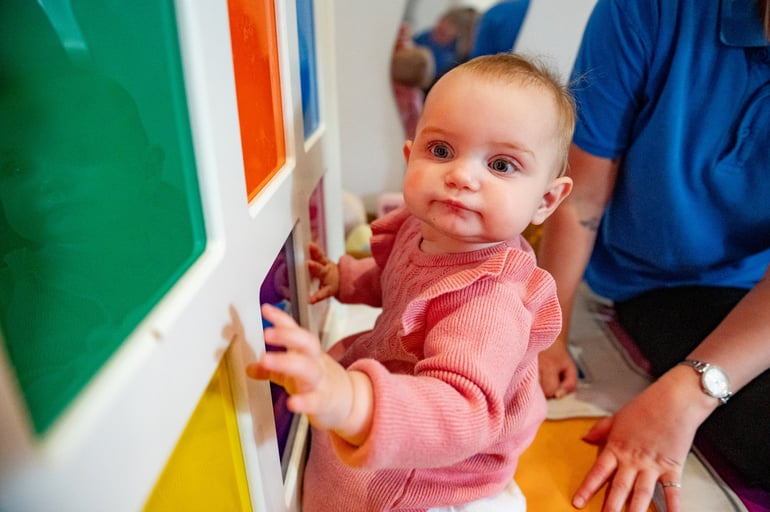Smiling baby in a pink sweater reaches toward colourful activity panels beside an adult, conveying curiosity and playful learning.