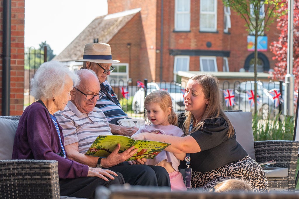 A family sits in a garden, reading a storybook to a young child, surrounded by greenery and flowers