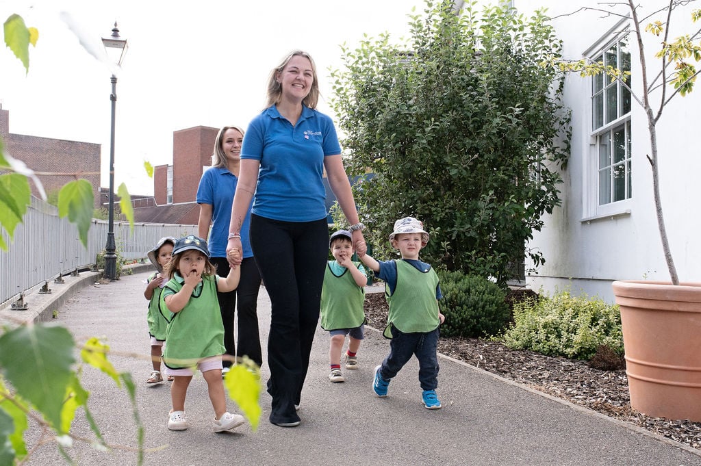 A woman walks with children along a sunny path in a nursery setting, surrounded by greenery and colourful flowers