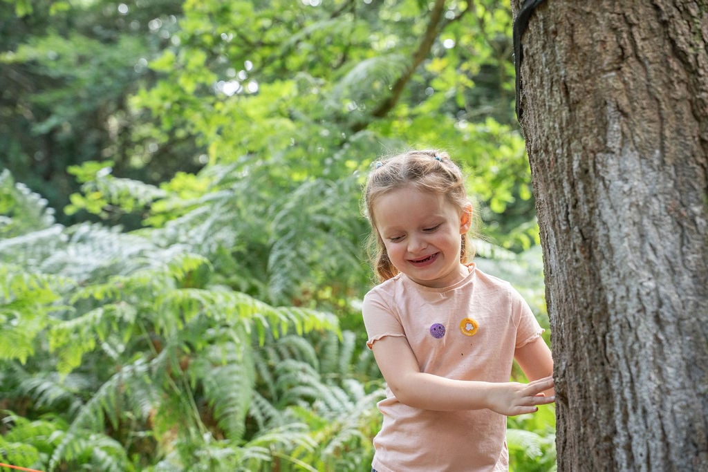A little girl stands beside a tree in a sunny nursery, smiling and enjoying the outdoors.
