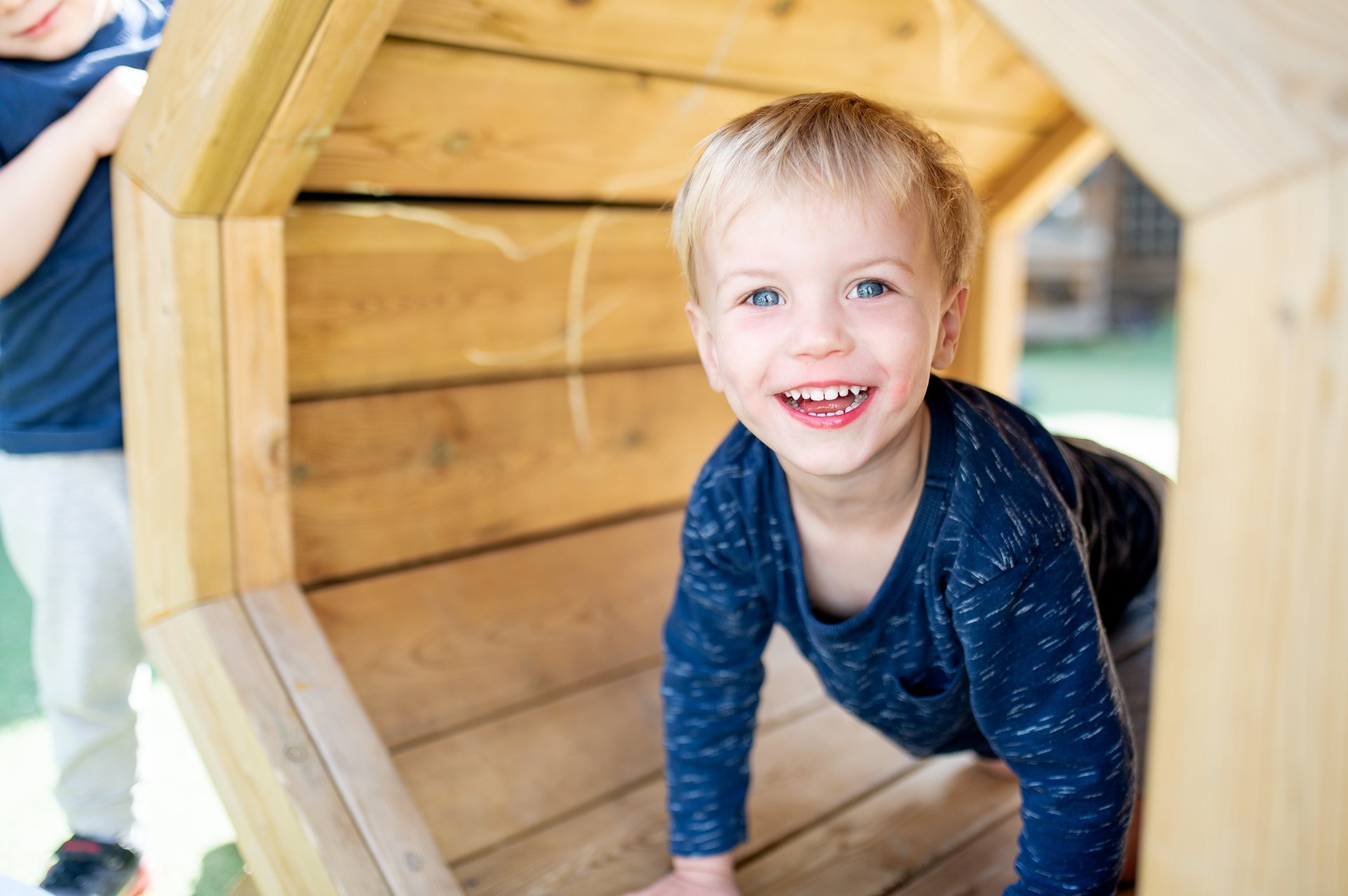 A young boy grins as he climbs into a wooden play structure in a nursery setting