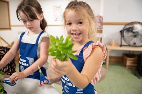 Two young girls in aprons happily holding green leaves in a nursery setting
