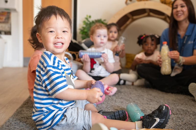 Smiling toddler in a blue-striped shirt holds a toy while diverse children play with a caregiver in a warm, colourful playroom.
