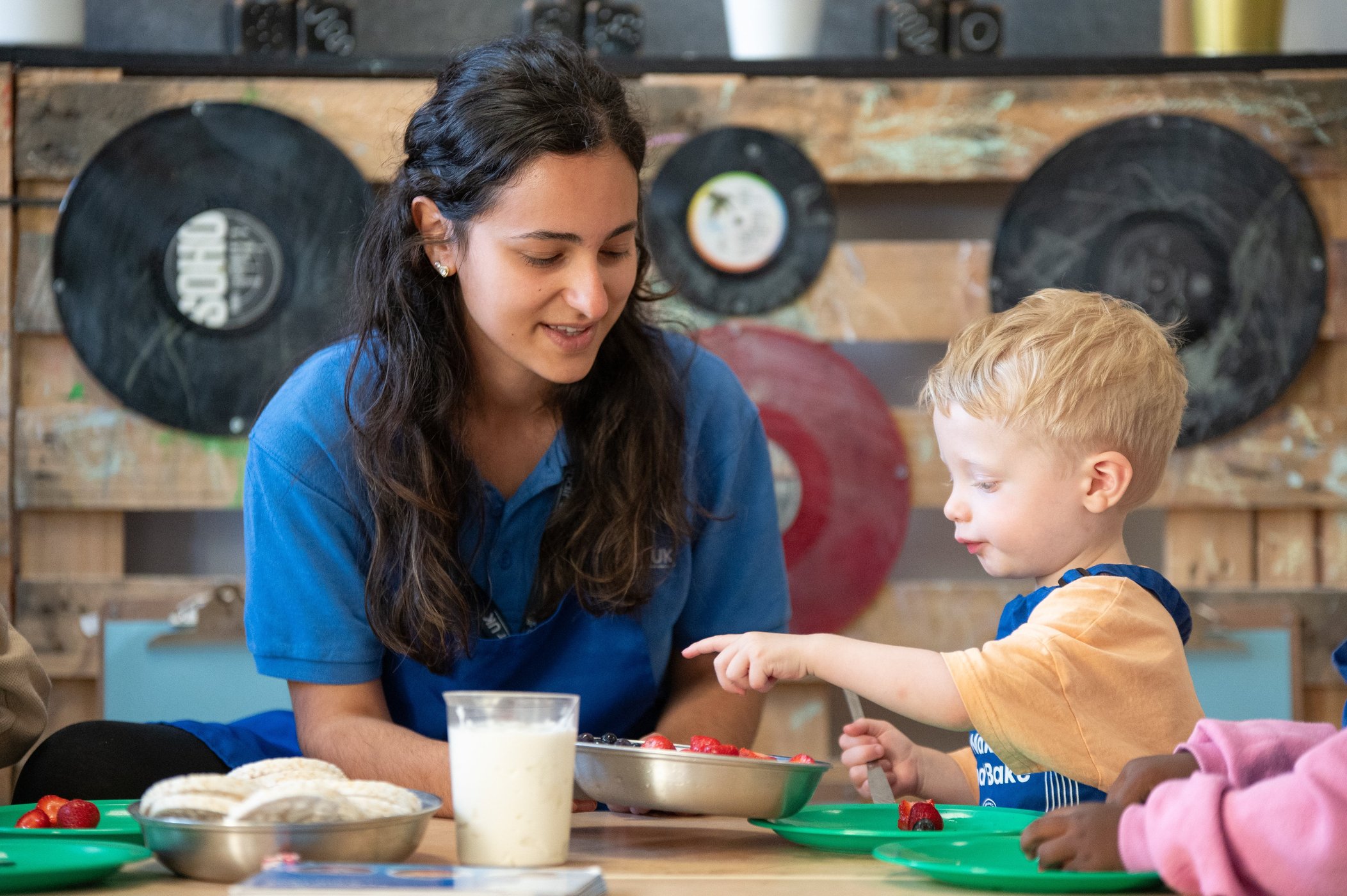 Smiling woman in blue helps a blond boy prepare food at a table; colourful bowls, milk, fruit, and vinyl records create a warm classroom scene.