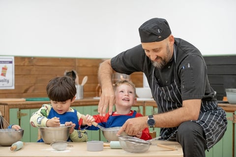 A man and two kids watch a chef cooking in a bright kitchen, filled with colourful ingredients and cheerful decor.