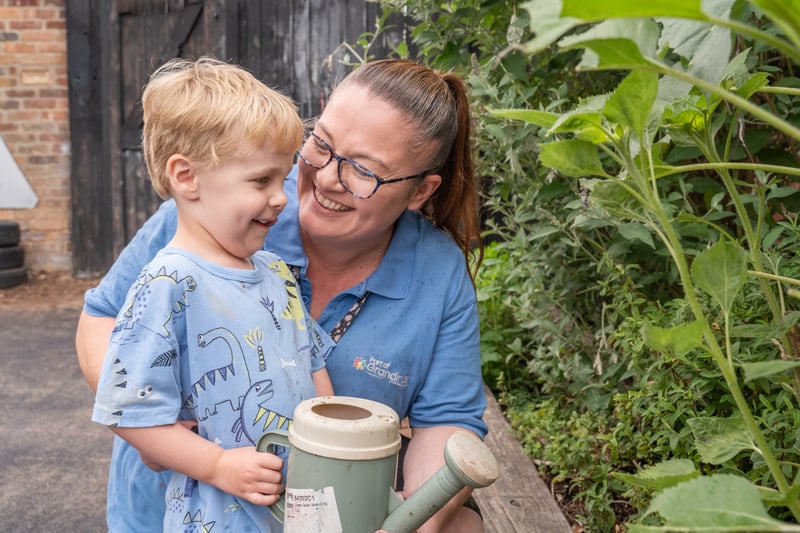 Smiling childcare worker hugs a young boy in a dinosaur shirt while holding a green watering can in a lush garden, conveying warmth and care.