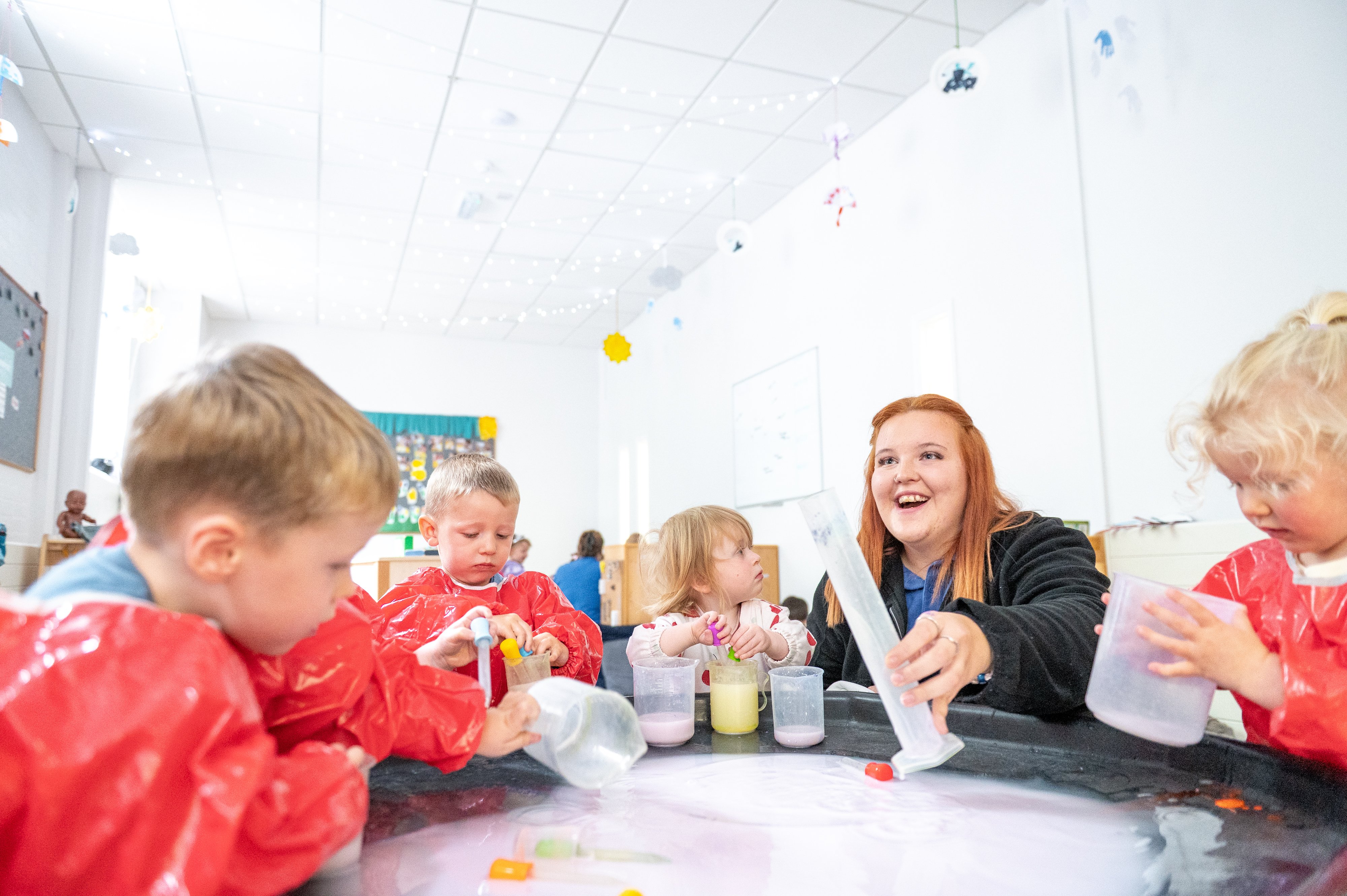 Children in red smocks explore colourful sensory materials at a classroom table while a smiling teacher guides them in a bright room.