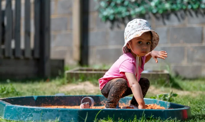 Child playing outside in a sand pit wearing a bucket hat at Kiddi Caru Day Nursery Preschool Abington Park Northampton