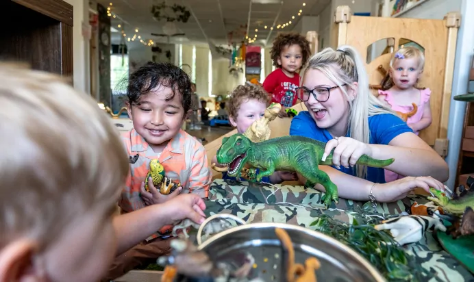 Key worker and children playing dinosaurs laughing at Kiddi Caru Day Nursery Preschool Abington Park Northampton