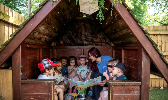 Key worker reading story to children in hut at Kiddi Caru Day Nursery Preschool Abington Park Northampton