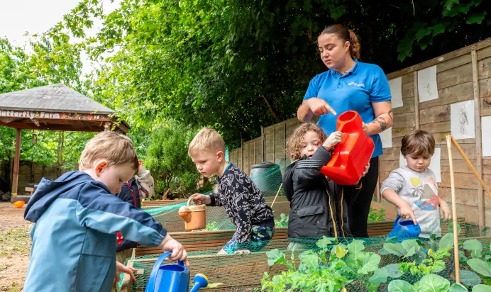 Key worker showing children how to water plants outside at Kiddi Caru Day Nursery Preschool Torquay