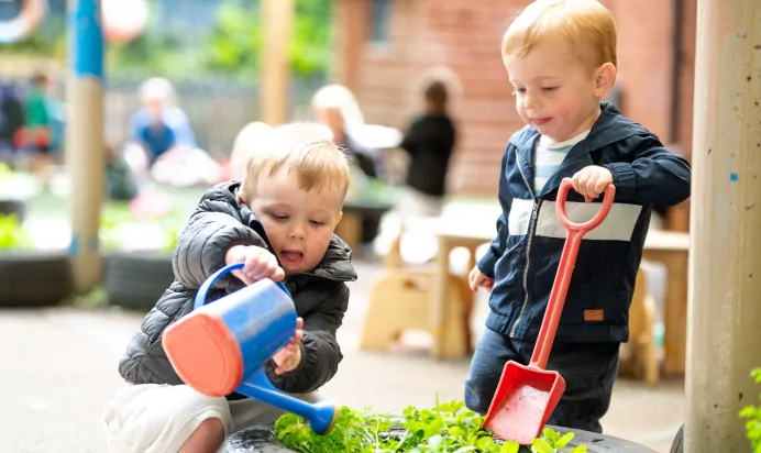 Two children watering a plant in the outdoor activity space at Kiddi Caru Day Nursery Preschool Torquay