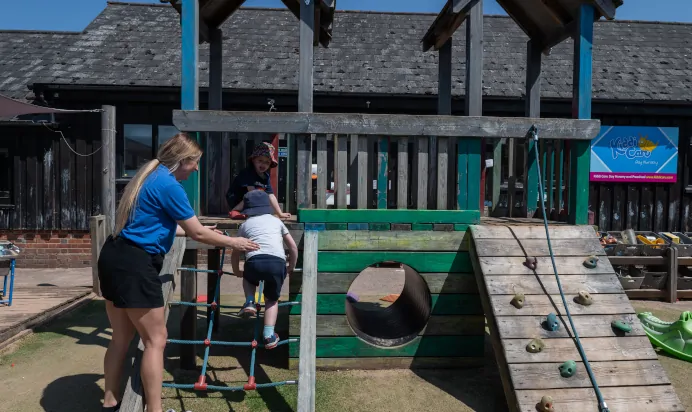Key worker helping child climb up rope ladder at Kiddi Caru Day Nursery Preschool Market Ower