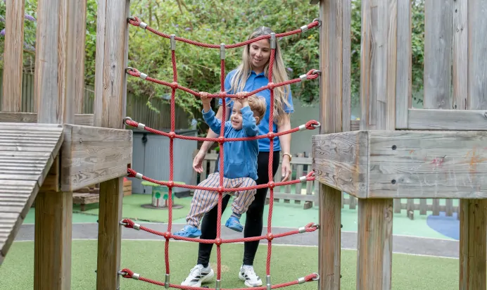 Key worker helping child climb on outdoor playground equipment at The Wandsworth Day Nursery Preschool London