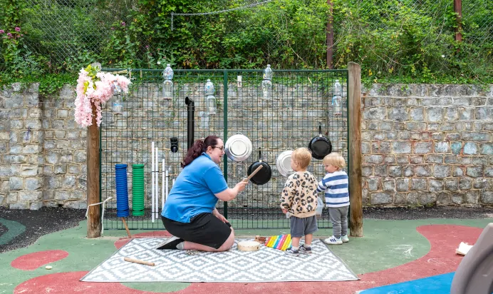 Key worker and children make music with pots in the garden at Kiddi Caru Day Nursery Preschool Crews Hole Bristol