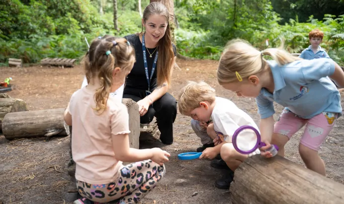 Children in woods using magnifying glass with key worker at The Puddleduck Preschool Priory Road Ascot