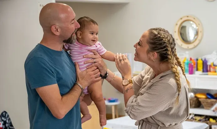 Man holding a baby, interacting with a woman in a home interior.