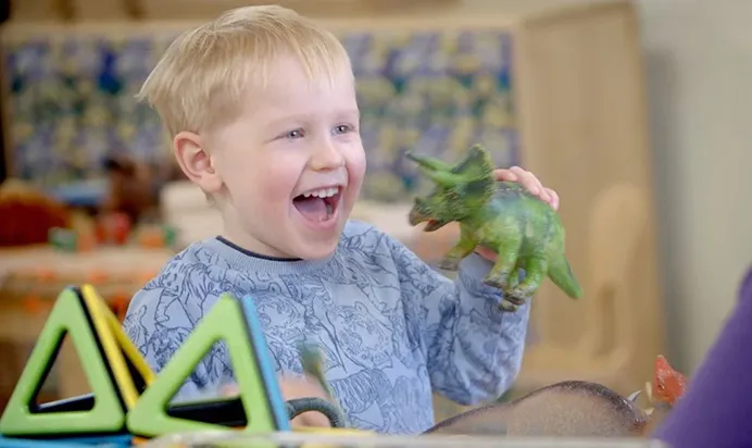 Child playing with a green dinosaur toy and colourful magnetic shapes on a table.