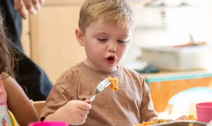 Child holding a fork with food, sitting at a table with a plate and cup visible.