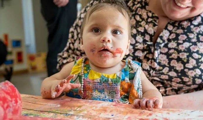A child wearing a colourful bib with paint-covered hands during an art activity; smiling adult beside them.