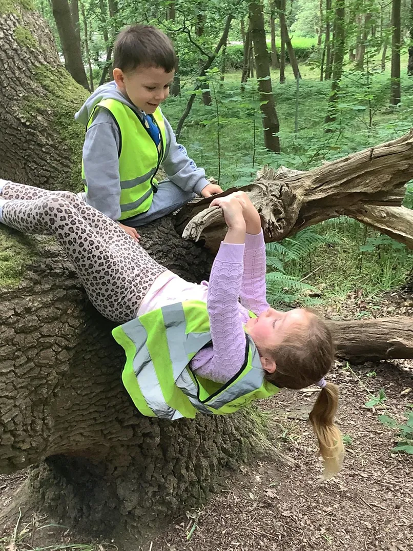 Two children in high-visibility vests playing on a fallen tree in a forest.