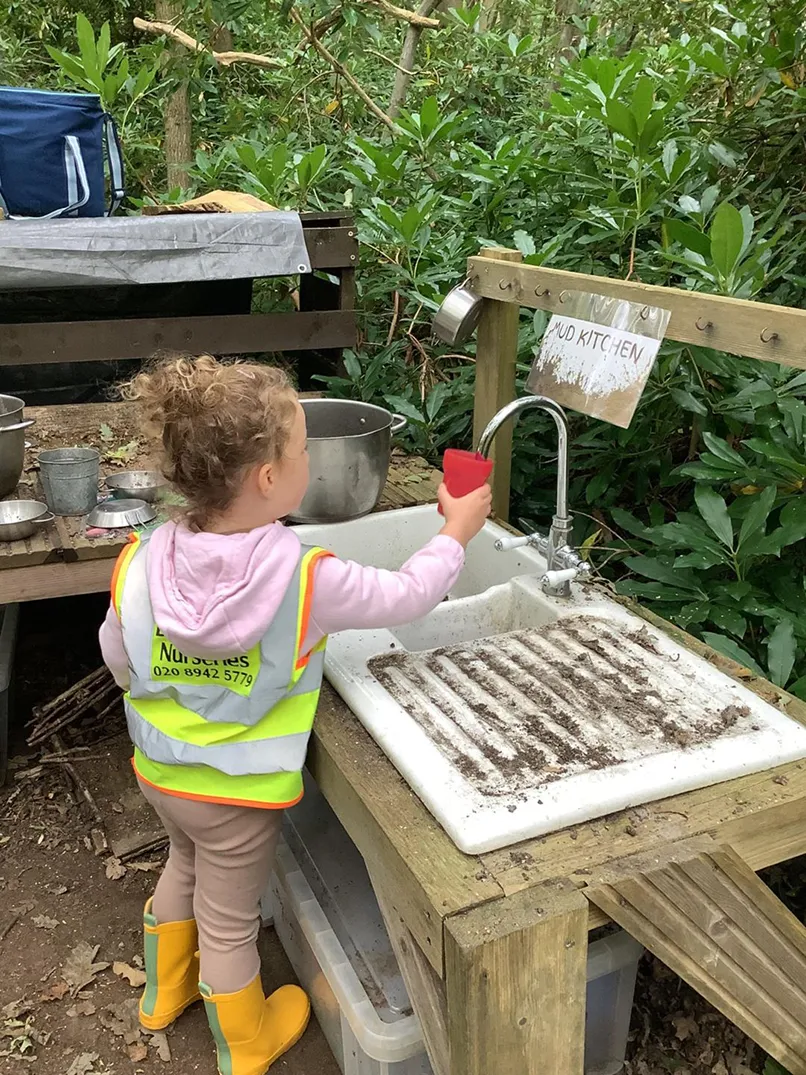 Child in a hi-vis vest playing at an outdoor mud kitchen with pots and sink.