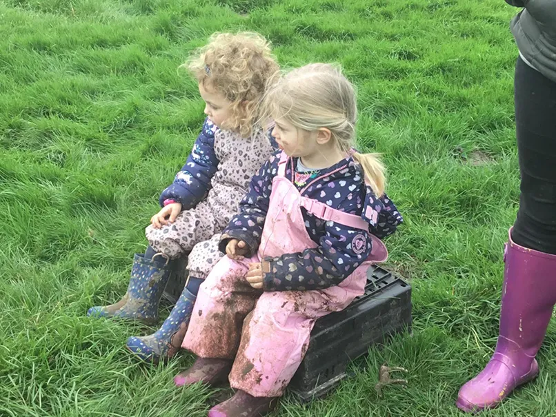 Two children in muddy clothes sitting on a crate in a grassy field, with a pair of purple boots visible.