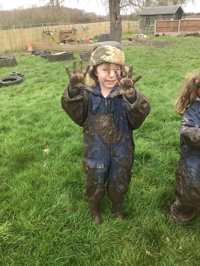 Child in a muddy overalls showing hands covered in mud, standing on grass with outdoor play area behind.