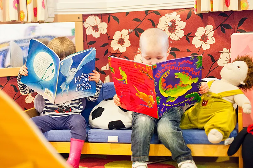 Two children reading books on a sofa with a colorful floral background and a doll beside them.
