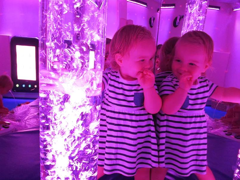 Child in striped outfit next to a glowing bubble lamp in a purple-lit sensory room.