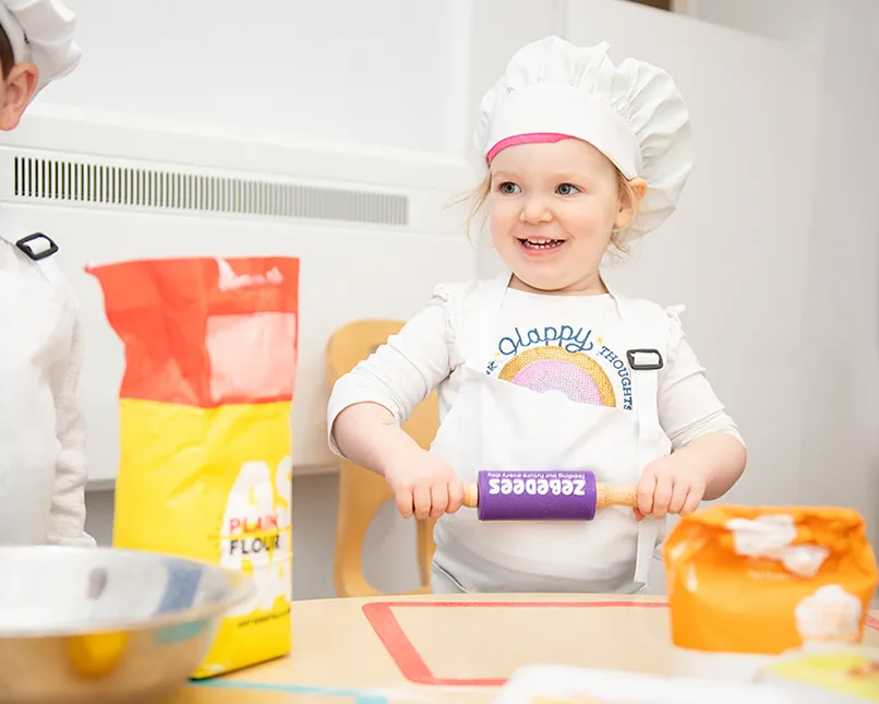 Child in a chef's hat with a rolling pin, next to flour bags and a mixing bowl.