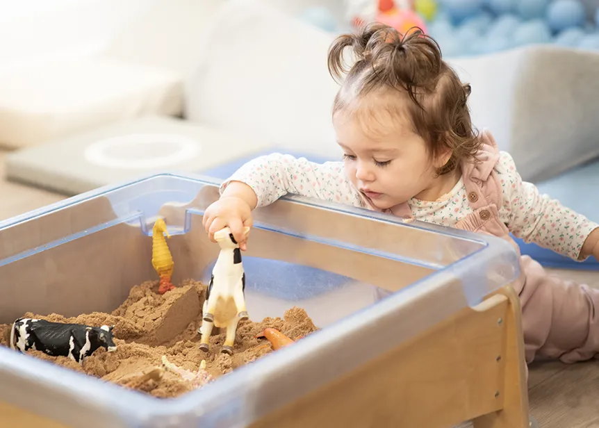 Child playing with toy animals in a sandbox indoors.