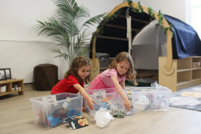 Two children sort waste into labelled bins for paper, general waste, and plastic in a colourful, playful environment.