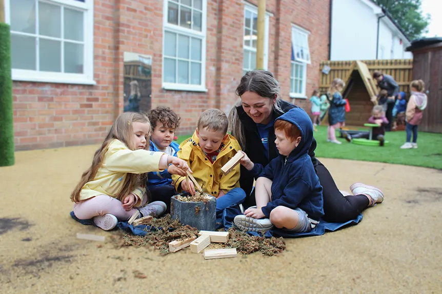 A group of children and an adult are engaged in a playful activity with wooden blocks and moss in an outdoor setting.
