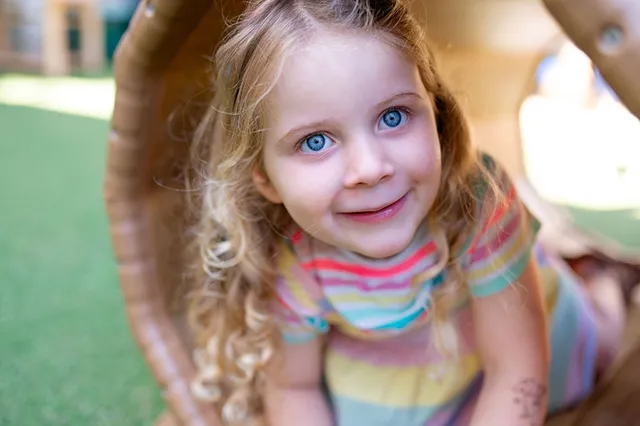 A child with curly hair crawls through a circular wooden tunnel, surrounded by green turf and playful outdoor scenery.