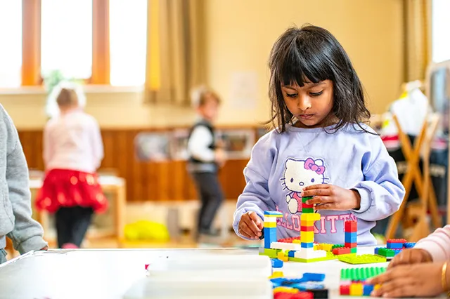 A child in a purple sweatshirt builds with colorful blocks on a table, engaged in play with other children in the background.