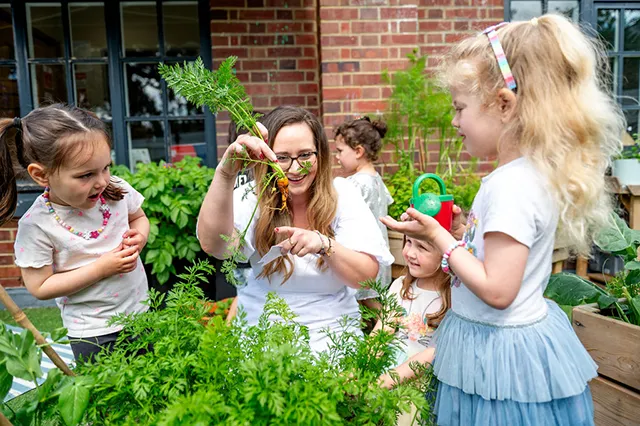 Children gather around a woman in a garden, marveling at freshly harvested carrots amidst lush greenery and gardening tools.