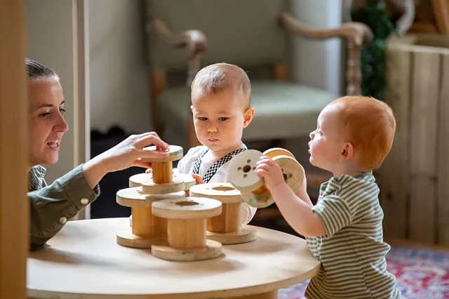 Two toddlers play with wooden toys on a table while an adult assists them in a cozy, softly lit room.