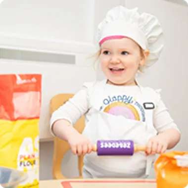 A child in a chef's hat and apron rolls out dough with a purple rolling pin, surrounded by bags of flour on a kitchen table.