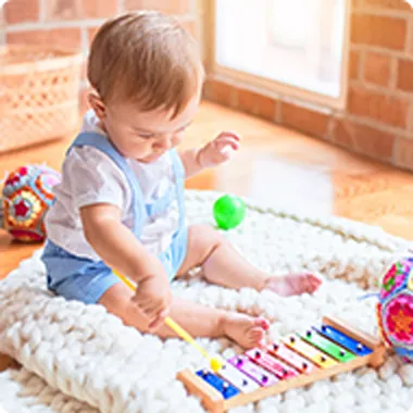 A toddler sits on a cosy knit blanket, happily playing a colorful xylophone, surrounded by vibrant balls and toys in a bright room.